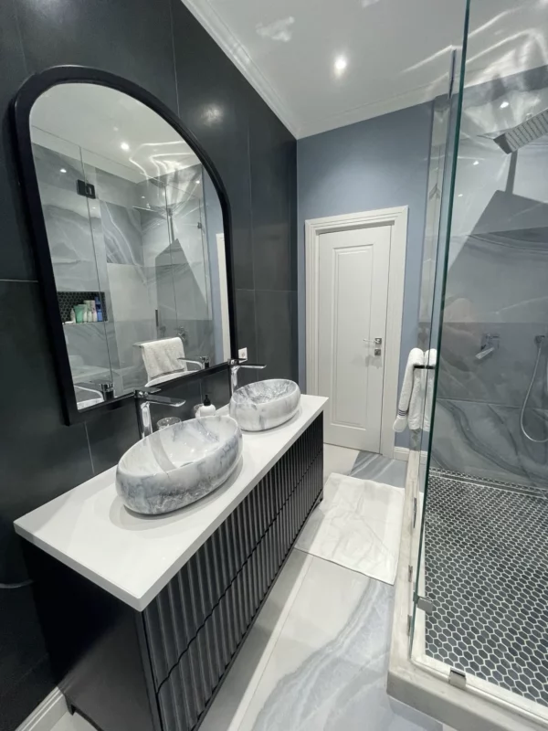 Bathroom remodel with two vessel sinks, a dark vanity, tall arched mirrors and a glass shower enclosure.