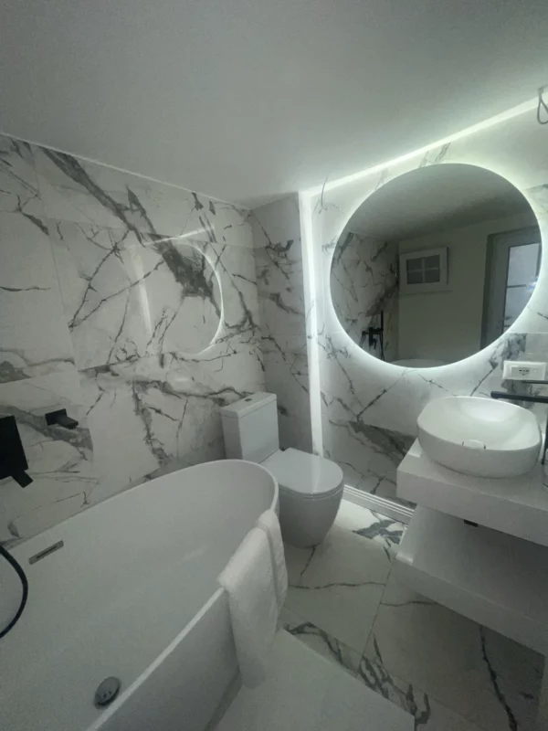 Bathroom remodel with a freestanding tub, veined wall and floor surfaces, a vessel sink and illuminated round mirror.