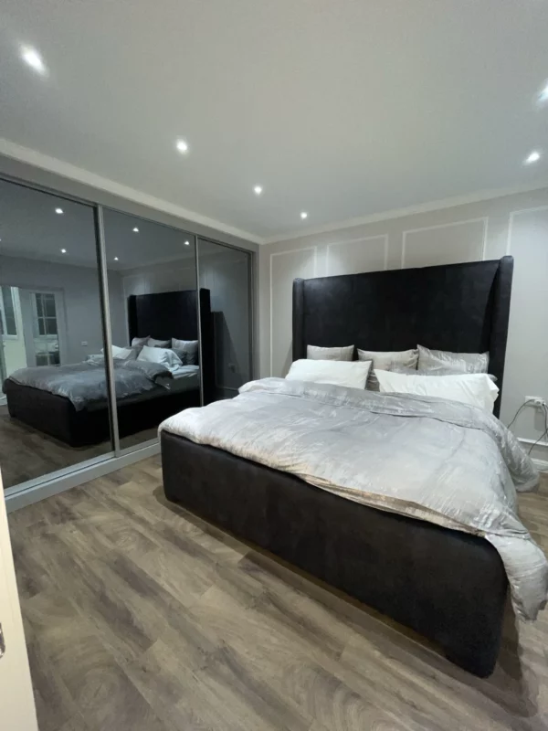 Bedroom in a whole-home renovation with a dark upholstered headboard, mirrored wardrobe doors, decorative wall molding and recessed lighting.
