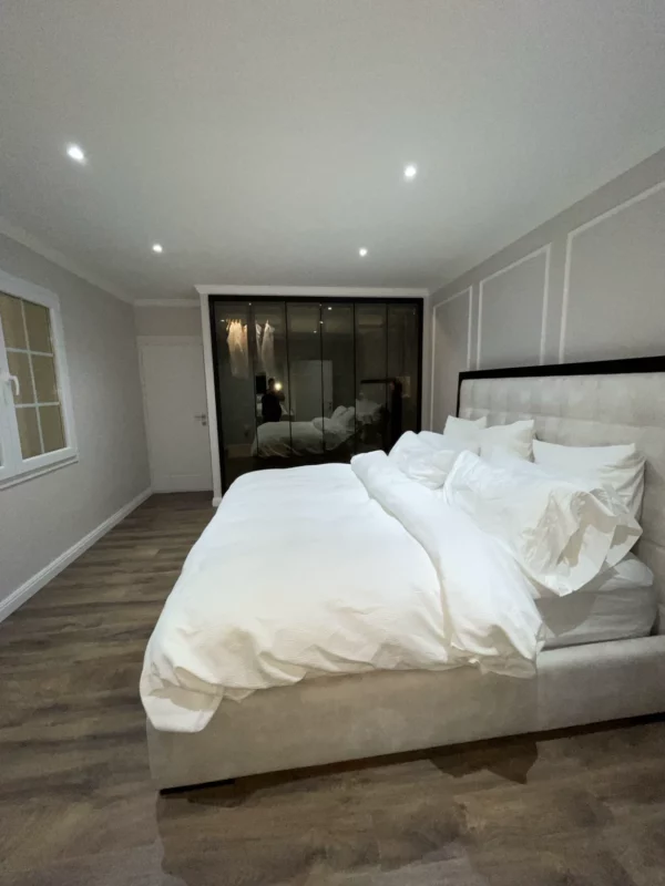 Second bedroom in the same whole-home renovation, with a light upholstered headboard, dark glass-front wardrobe, decorative wall molding and recessed lighting.