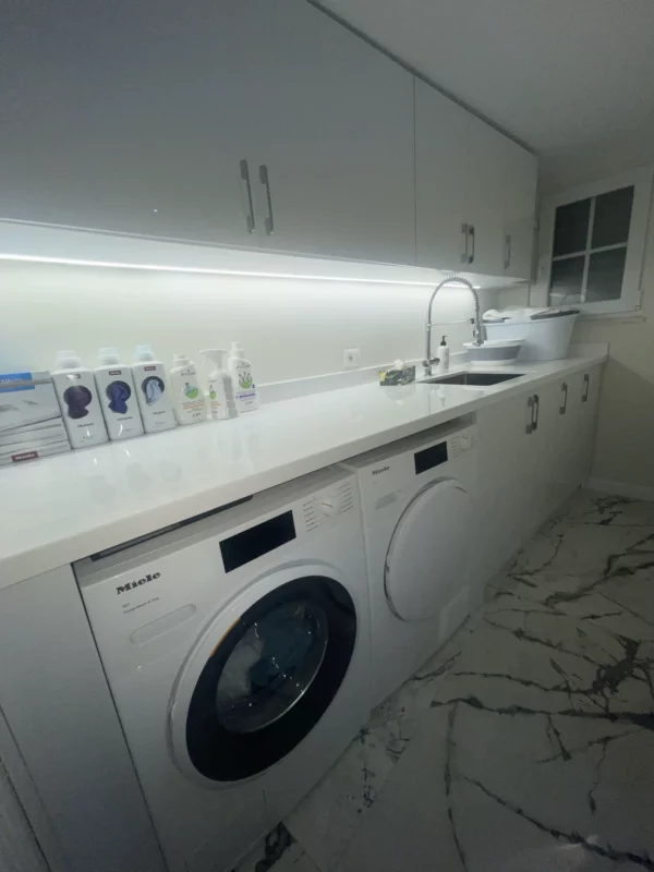 Laundry room remodel with front-loading appliances, white cabinetry, a long counter, sink and under-cabinet lighting.