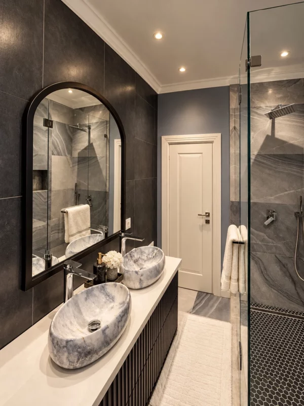 Double stone vessel basins, arched mirror, dark vanity and glass shower enclosure.